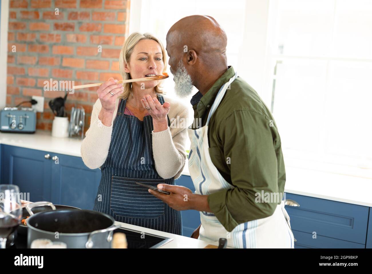 Glückliches, vielseitiges Paar in der Küche, das Schürzen trägt, zusammen kocht, Essen probiert Stockfoto