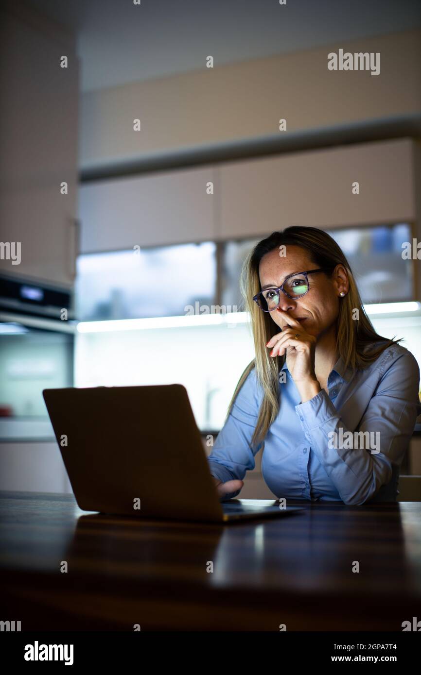 Hübsche Frau mittleren Alters, die spät am Tag auf einem Laptop-Computer zu Hause arbeitet, ein Geschäft von zu Hause aus führt, aus der Ferne arbeitet - frustriert wird Stockfoto