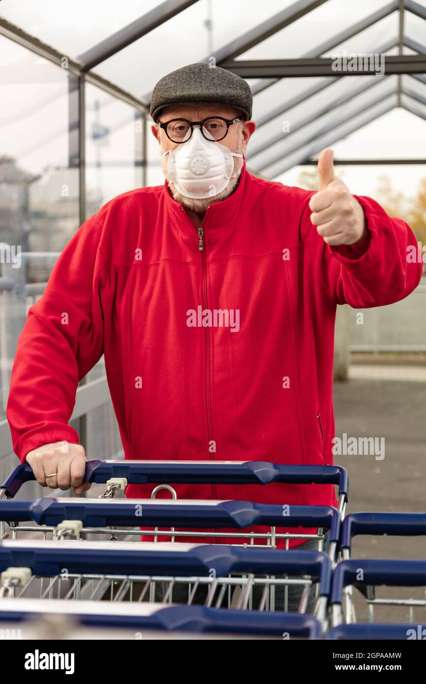Ein älterer Mann trägt eine N95-Atemschutzmaske, während er einen Einkaufswagen außerhalb eines Lebensmittelladens schiebt. Der Mann schützt sich vor Coronavirus und Stockfoto