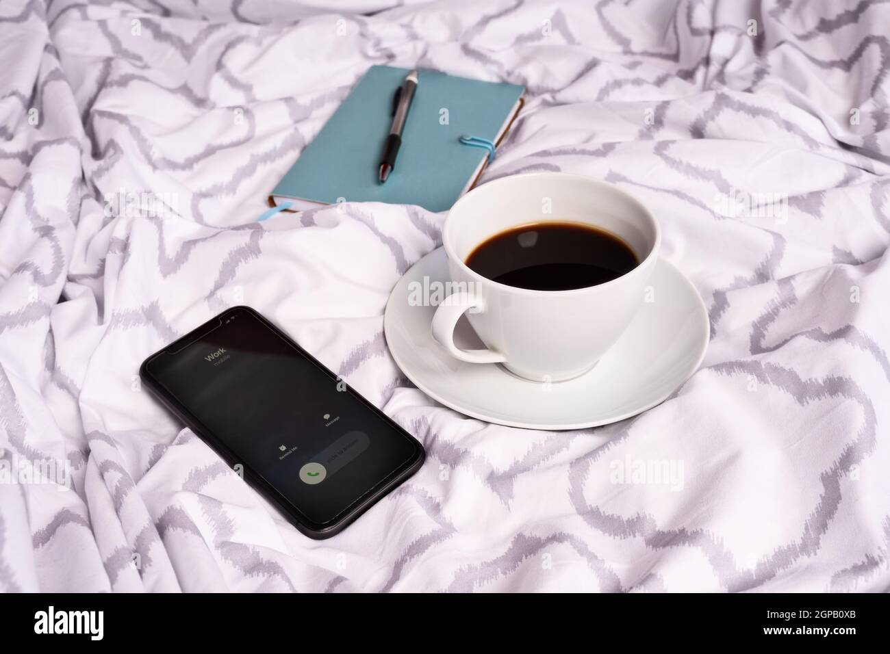 Tasse Kaffee auf modernen Bettlaken, Notebook-Stift und ein ankommender Anruf von der Arbeit auf einem Handy. Freiberufliches Konzept. Entgangene Anrufe von der Arbeit. Stockfoto