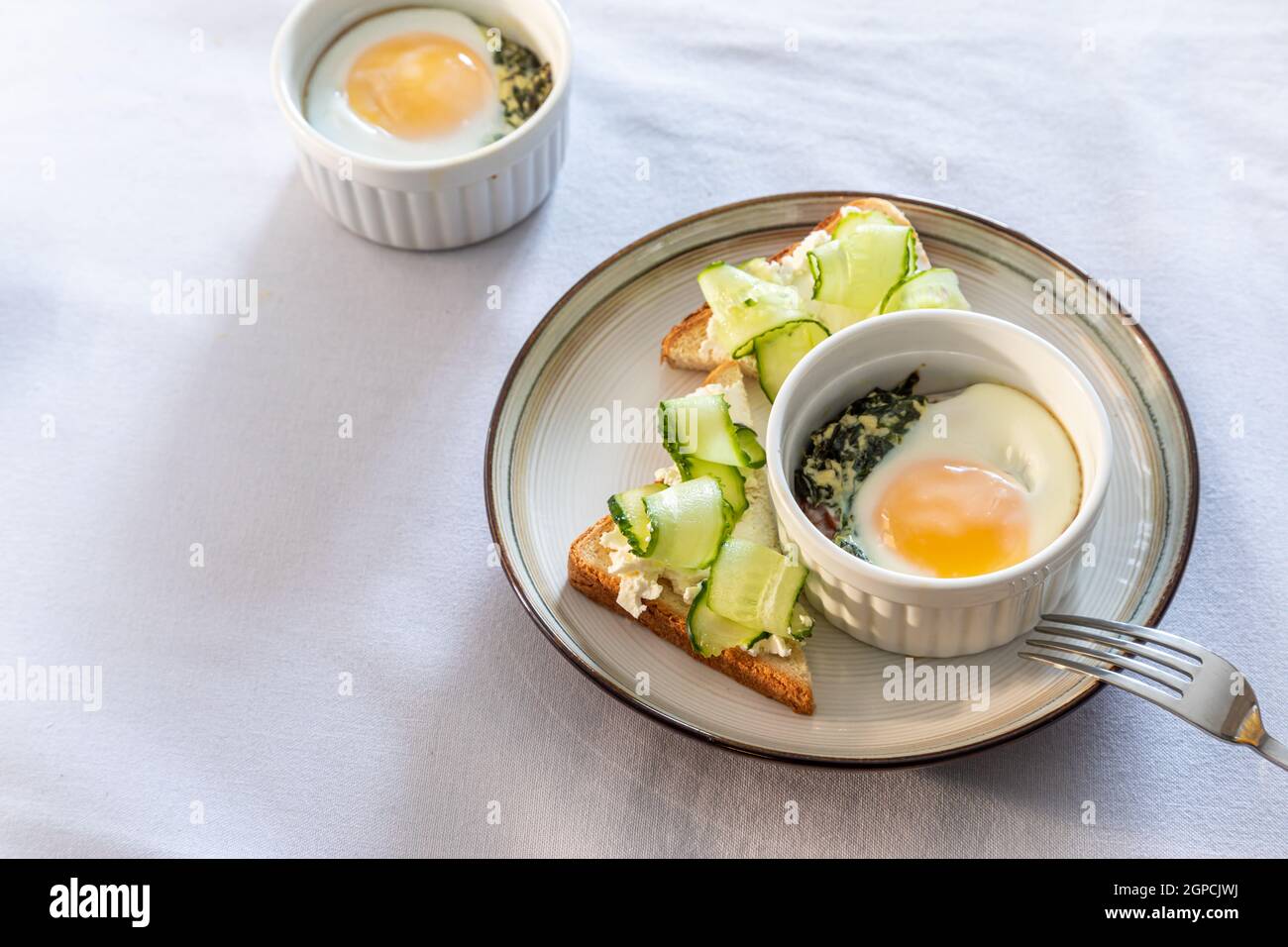Geraffte Eier (Oeuf Cocotte) oder gebackene Eier. Gesundes Frühstück mit Eiern und Spinat und Toast mit Gurke Stockfoto