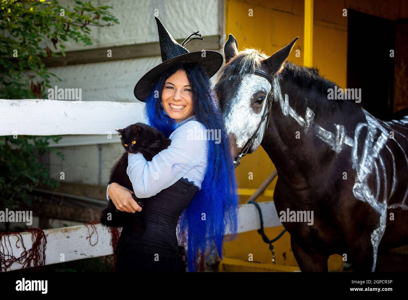 Ein Mädchen, das als Hexe gekleidet ist, hält eine schwarze Katze In ihren Armen und steht bei einem Corral auf einem Farm neben einem Pferd Stockfoto