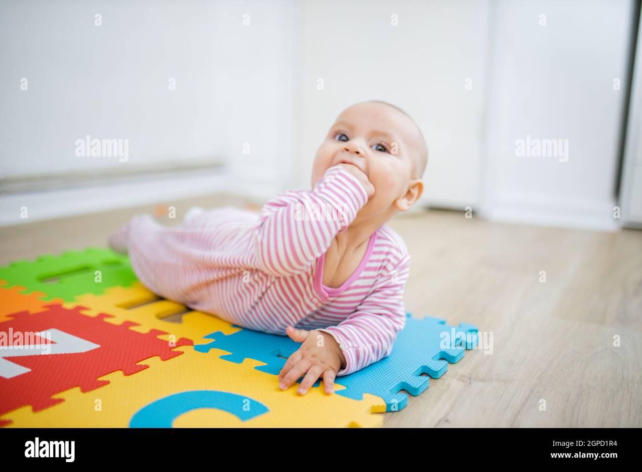 Entzückendes Baby, das mit dem Gesicht nach unten auf der bunten Kindermatte liegt, mit der Faust im Mund. Portrait von fröhlichem Baby, das auf dem Boden spielt. Glückliche Babys mit f Stockfoto