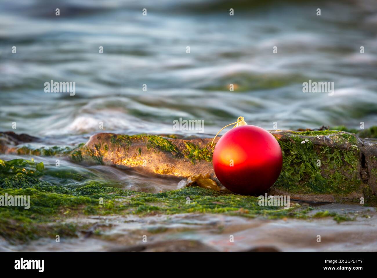 Ein australisches weihnachtsfest am Wasser, eine rote Kugel am Strand und Felsen bei Sonnenuntergang mit Wellen, die den warmen Abendhimmel reflektieren Stockfoto