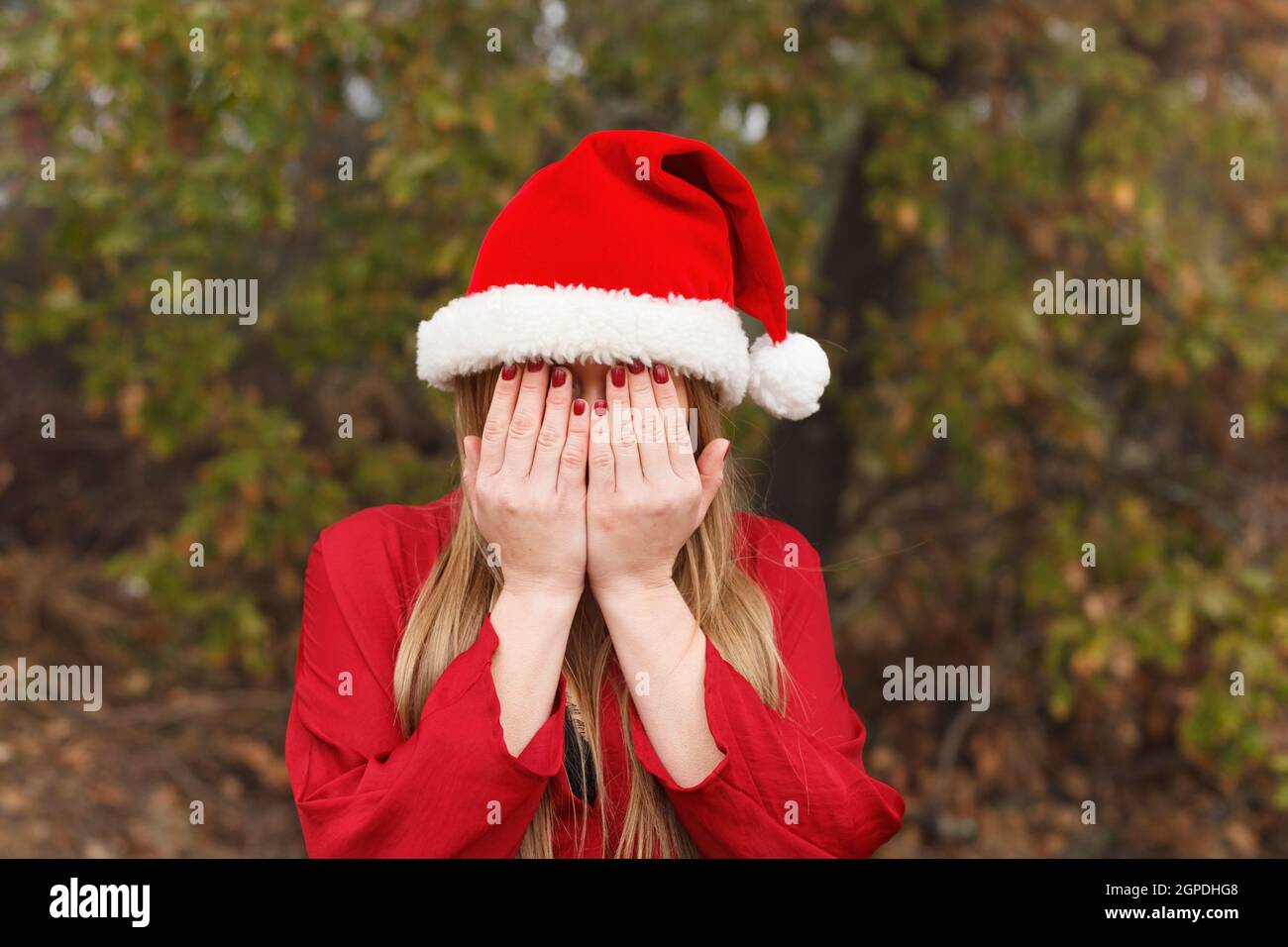 Blonde Frau mit Weihnachtshut, die ihr Gesicht in einem Park bedeckt Stockfoto