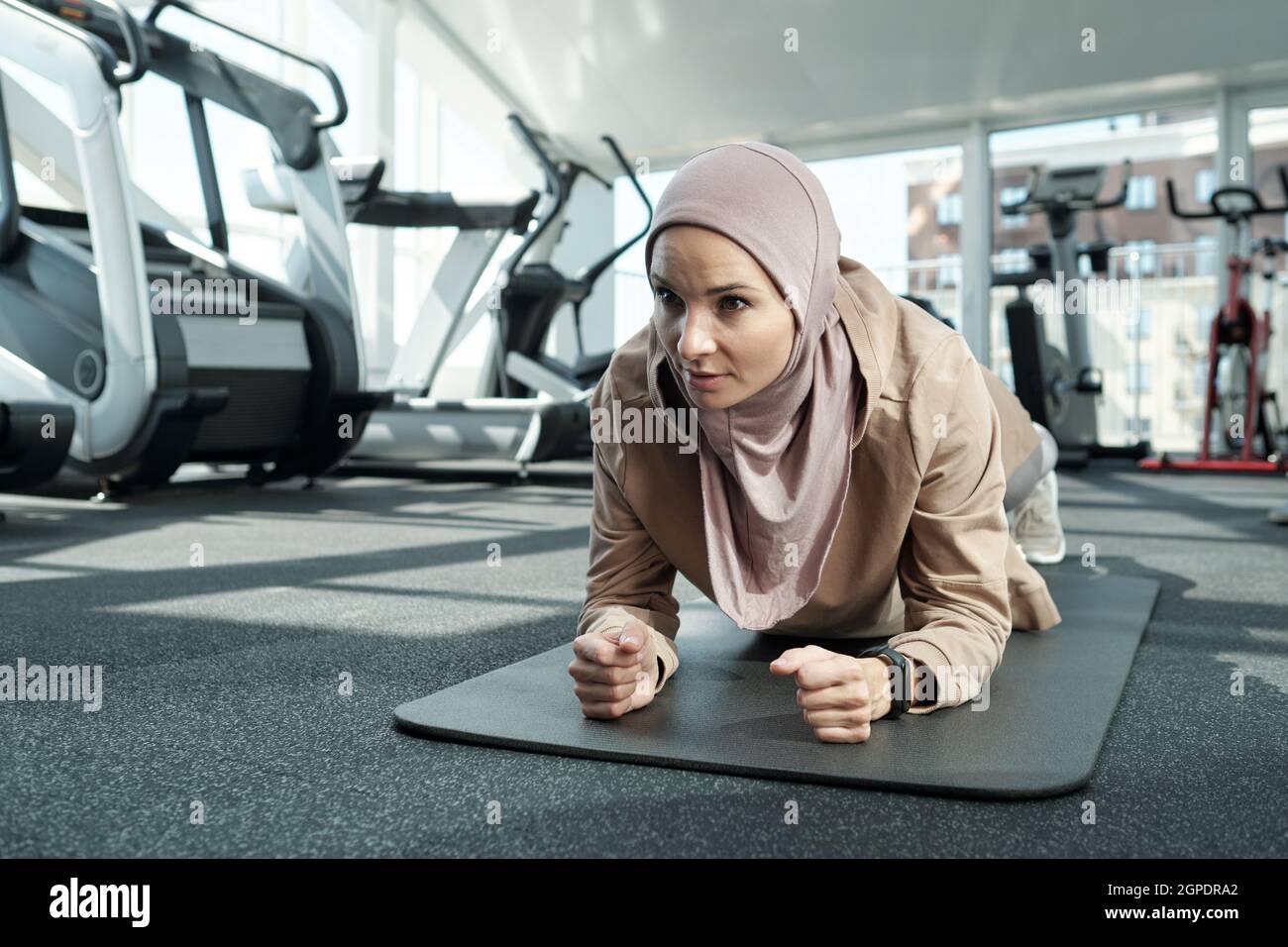 Horizontale, mittlere Nahaufnahme einer starken jungen, erwachsenen muslimischen Frau, die Hijab trägt und im Fitnessstudio Ellenbogenplankenübungen macht Stockfoto