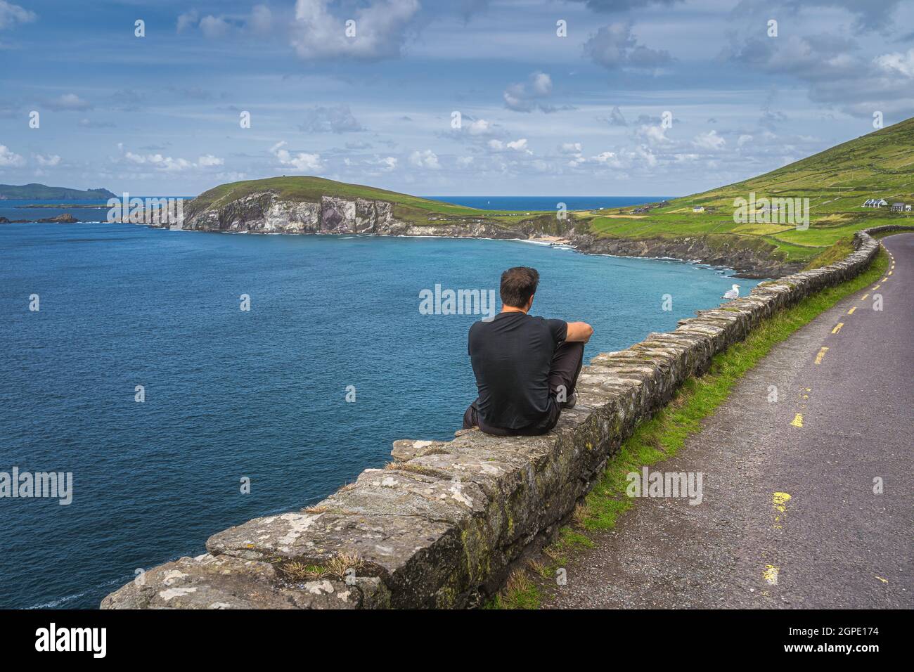 Mann mittleren Alters, der an einer Wand sitzt und den atemberaubenden Blick auf Slea Head, Coumeenoole Beach und die Dingle Peninsula, Teil des Wild Atlantic Way, Kerry, Irland, bewundert Stockfoto