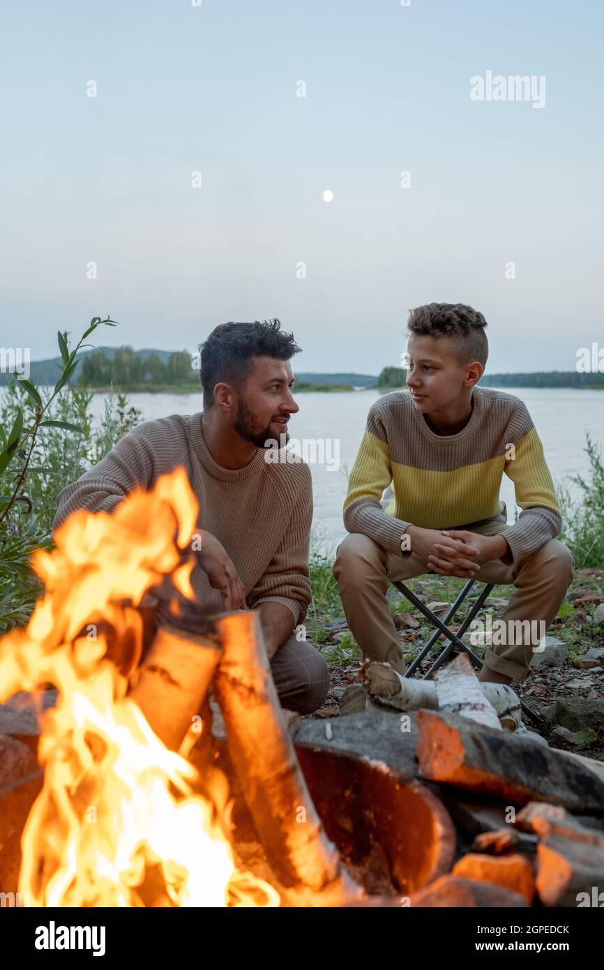 Junger Vater und Sohn sitzen am Lagerfeuer auf dem Hintergrund des Sees am Abend Stockfoto