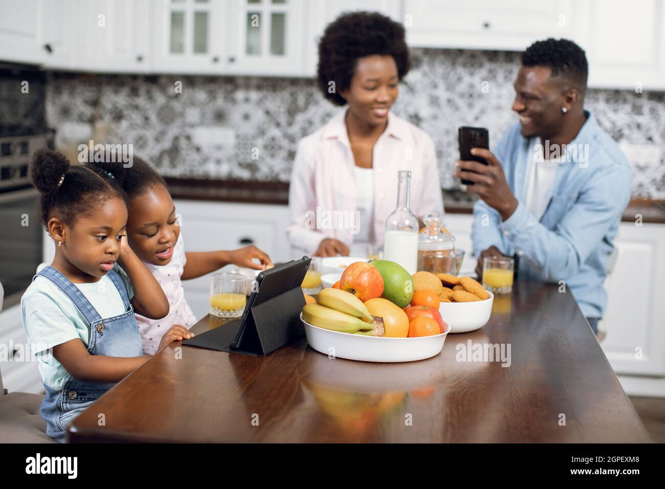 afroamerikanische Eltern sitzen in der Küche und nutzen das Smartphone, während ihre niedlichen Töchter Zeichentrickfilme auf dem digitalen Tablet ansehen. Moderne Gadgets für den Lebensstil. Morgens zu Hause. Stockfoto