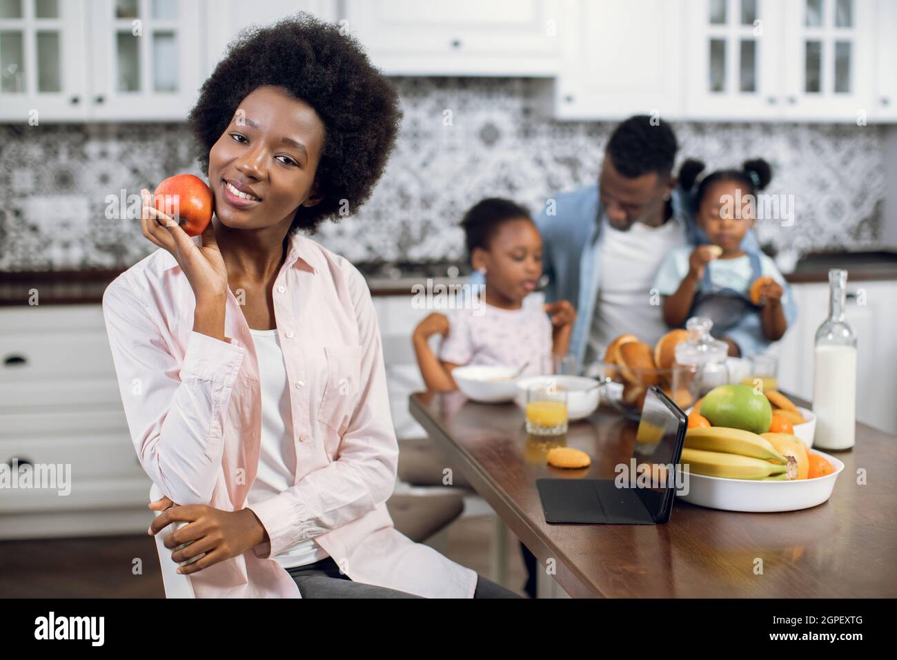Angenehme afroamerikanische Frau, die lächelt und die Kamera anschaut, während sie in der Küche mit Apfel in den Händen sitzt. Ehemann und zwei Töchter spielen im Hintergrund. Häusliches Leben der glücklichen Familie. Stockfoto