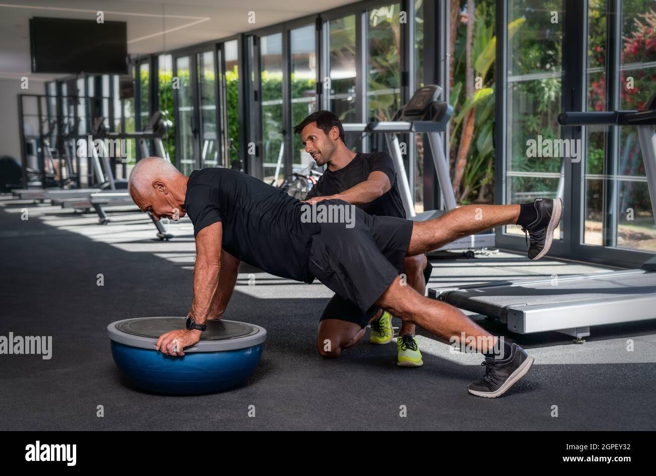Ein älterer kaukasischer Mann, im Fitnessstudio, mit einem persönlichen Fitnesstrainer, in der Rehabilitation, macht Übungen für das Gleichgewicht auf einem Gymnastikball. Stockfoto