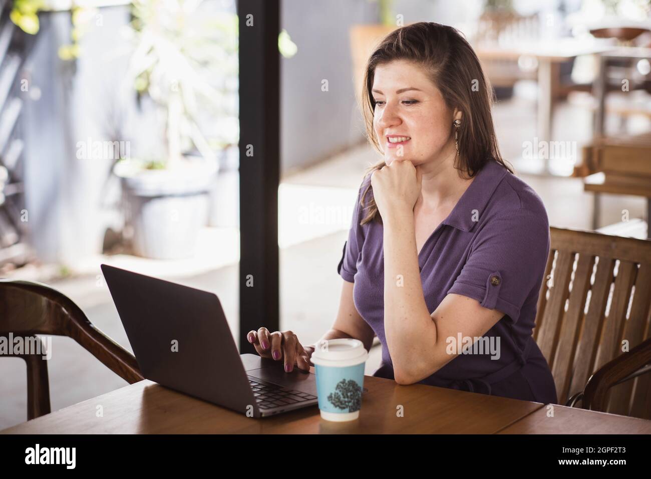 Porträt einer schönen Frau, die am Laptop arbeitet, während sie im Café mit Kaffee in einer Papiertasse sitzt. Sie hat ein Online-Meeting. Konzept funktioniert in Innenräumen Stockfoto