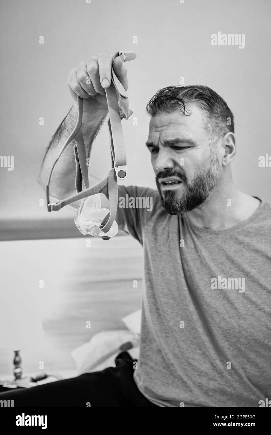 Unglücklich schockiert Mann mit chronischen Atemproblemen überrascht, indem CPAP-Maschine auf dem Bett im Schlafzimmer sitzen. Stockfoto