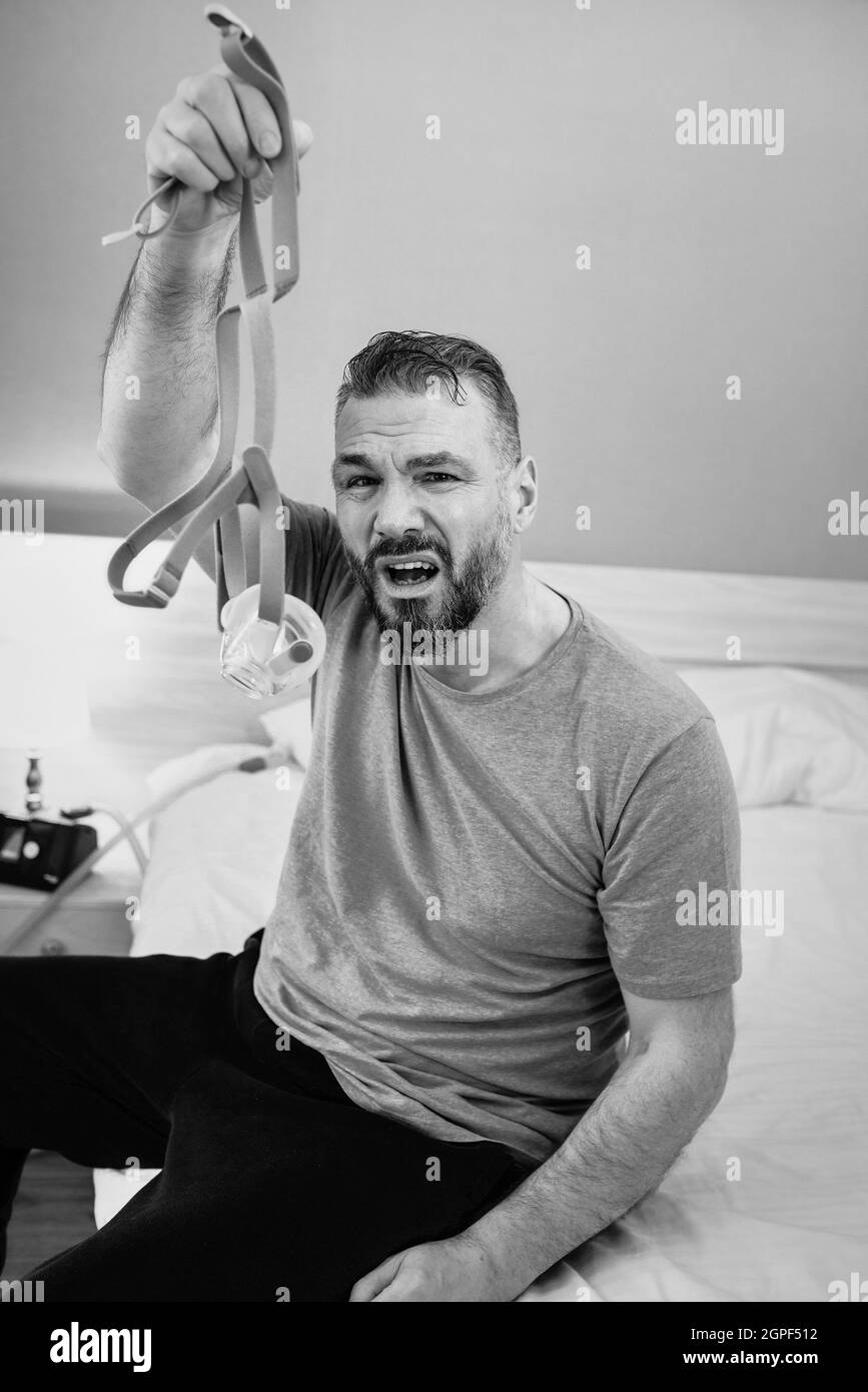 Unglücklich schockiert Mann mit chronischen Atemproblemen überrascht, indem CPAP-Maschine auf dem Bett im Schlafzimmer sitzen. Stockfoto