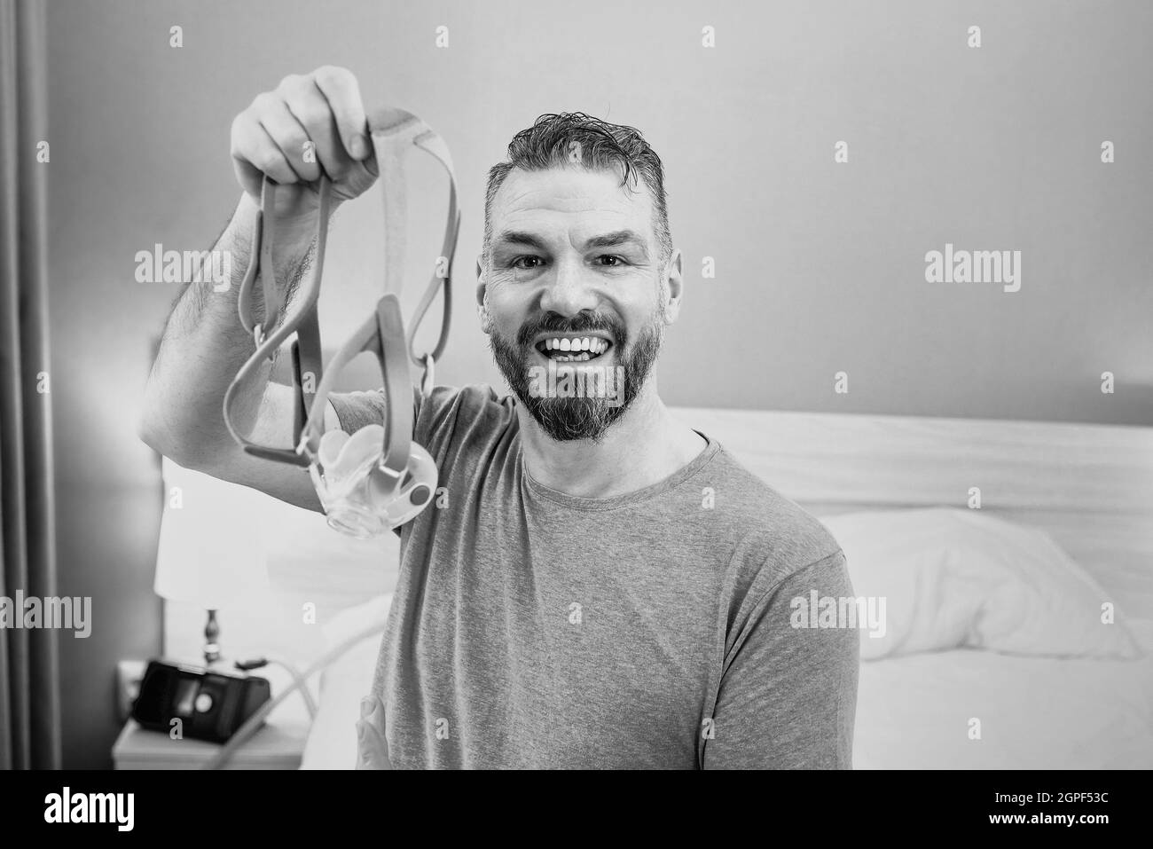 Monochromes Porträt eines glücklichen ausgeruhten Mannes mit chronischen Atemproblemen nach der Verwendung eines CPAP-Geräts, das im Schlafzimmer auf dem Bett sitzt. Gesundheitswesen, CPAP Stockfoto