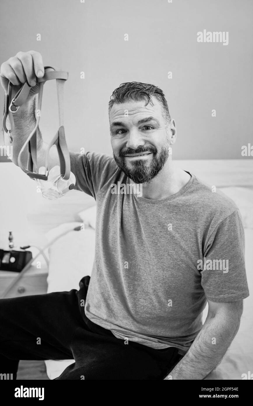 Monochromes Porträt eines glücklichen ausgeruhten Mannes mit chronischen Atemproblemen nach der Verwendung eines CPAP-Geräts, das im Schlafzimmer auf dem Bett sitzt. Gesundheitswesen, CPAP Stockfoto