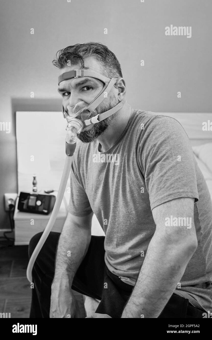 Unglücklich schockiert Mann mit chronischen Atemproblemen überrascht, indem CPAP-Maschine auf dem Bett im Schlafzimmer sitzen. Stockfoto