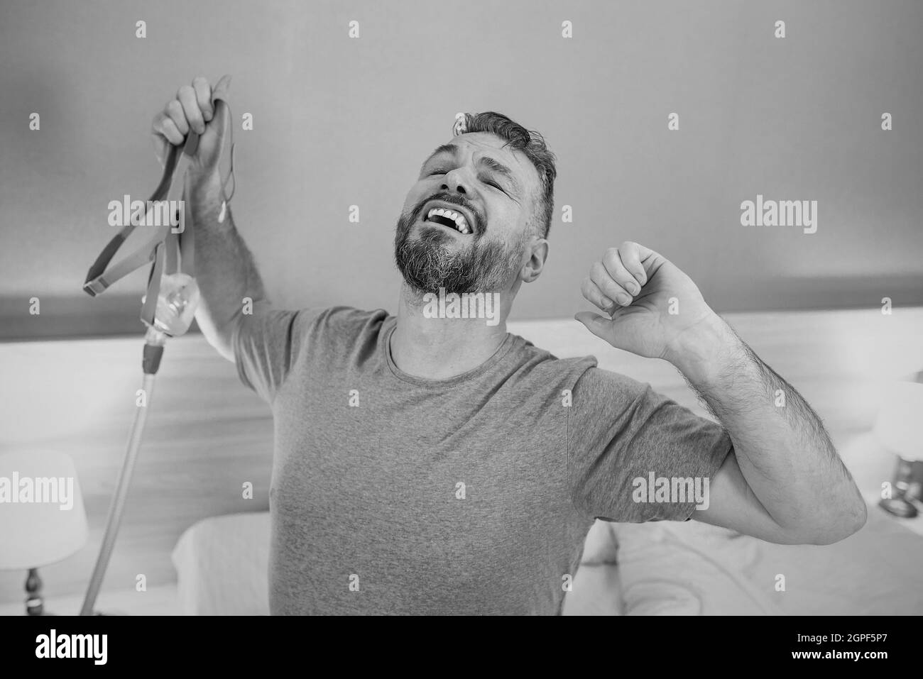 Monochromes Porträt eines glücklichen ausgeruhten Mannes mit chronischen Atemproblemen nach der Verwendung eines CPAP-Geräts, das im Schlafzimmer auf dem Bett sitzt. Gesundheitswesen, CPAP Stockfoto