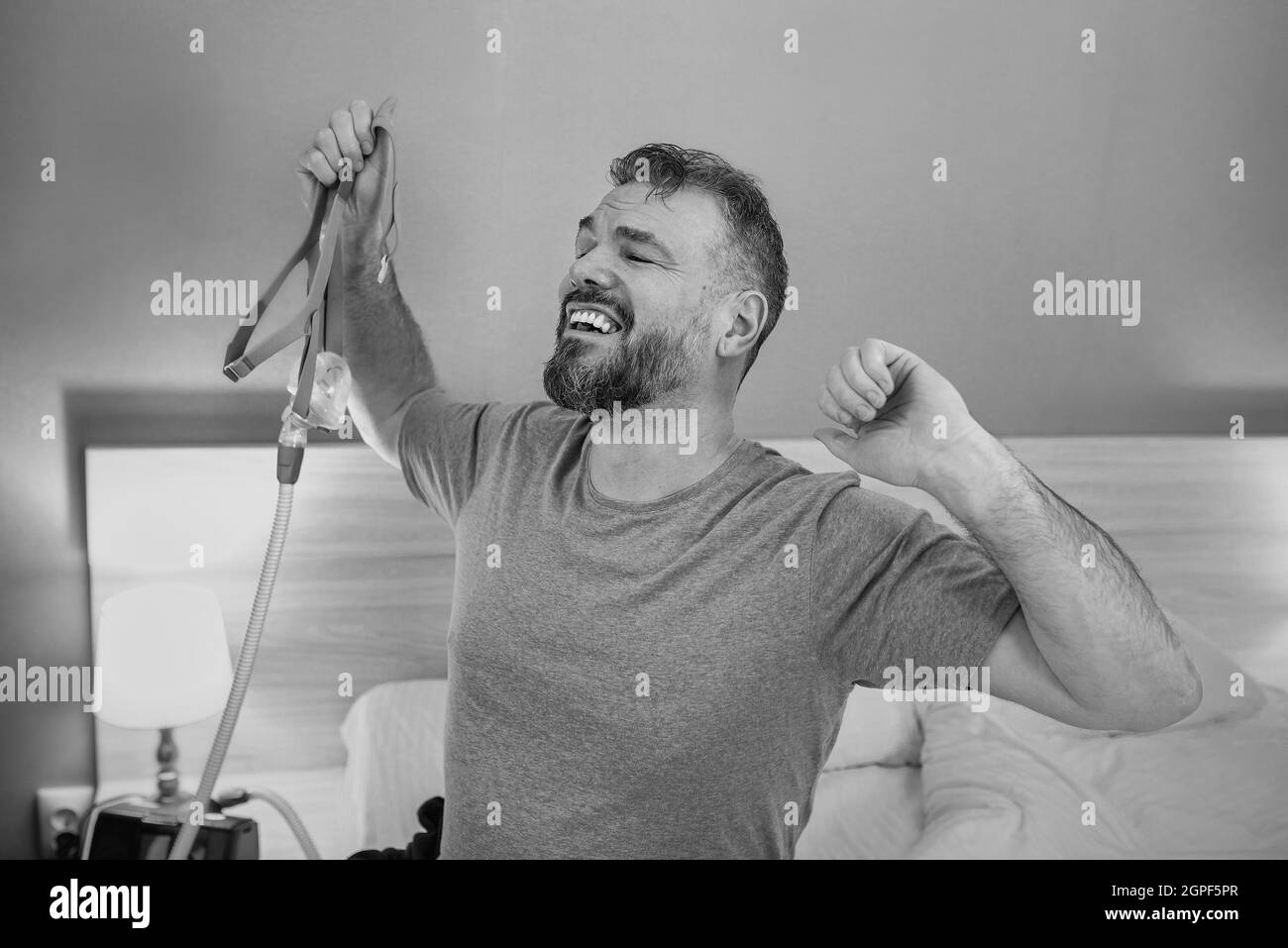 Monochromes Porträt eines glücklichen ausgeruhten Mannes mit chronischen Atemproblemen nach der Verwendung eines CPAP-Geräts, das im Schlafzimmer auf dem Bett sitzt. Gesundheitswesen, CPAP Stockfoto