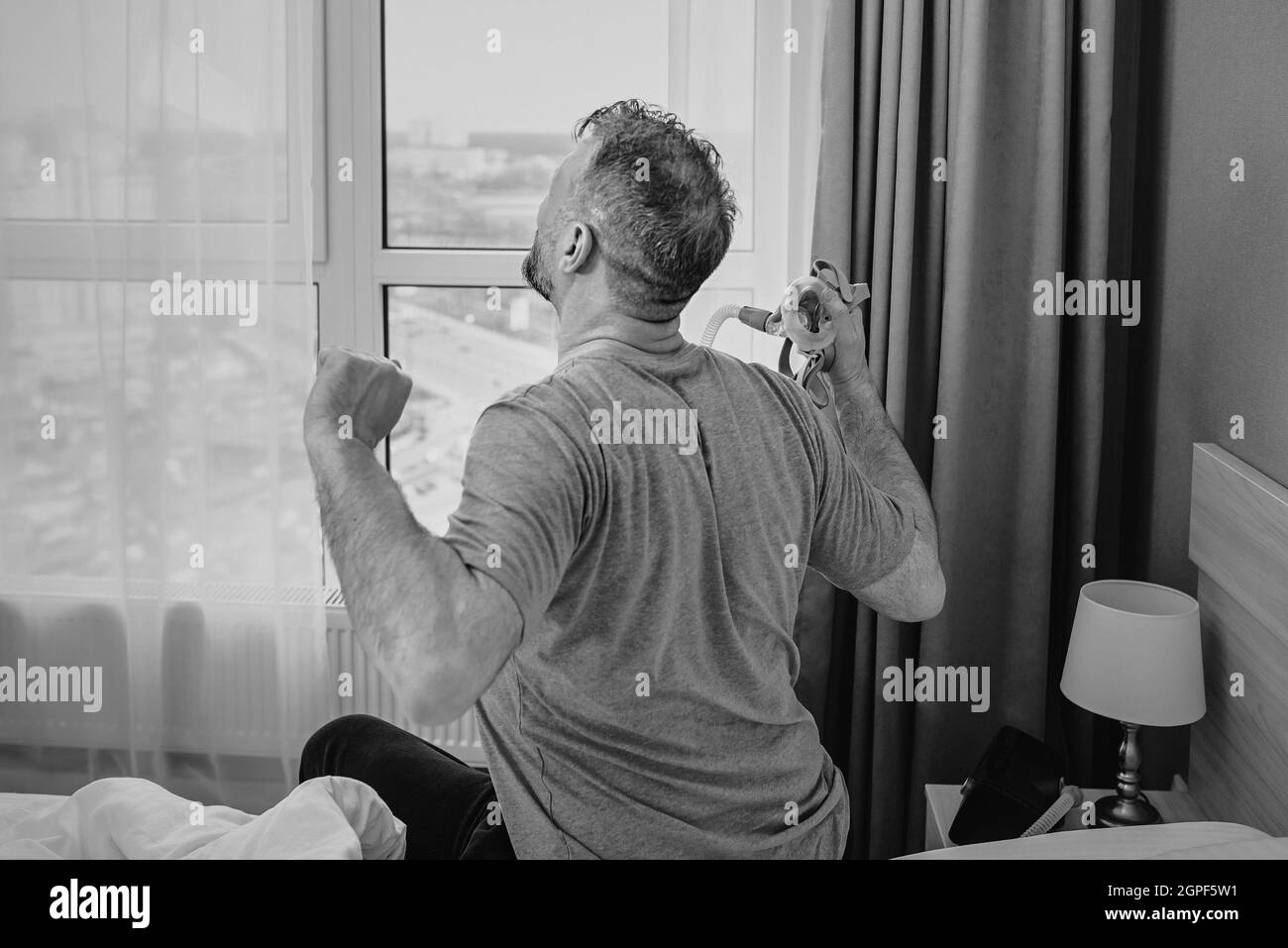 Monochromes Porträt eines glücklichen ausgeruhten Mannes mit chronischen Atemproblemen nach der Verwendung eines CPAP-Geräts, das im Schlafzimmer auf dem Bett sitzt. Gesundheitswesen, CPAP Stockfoto