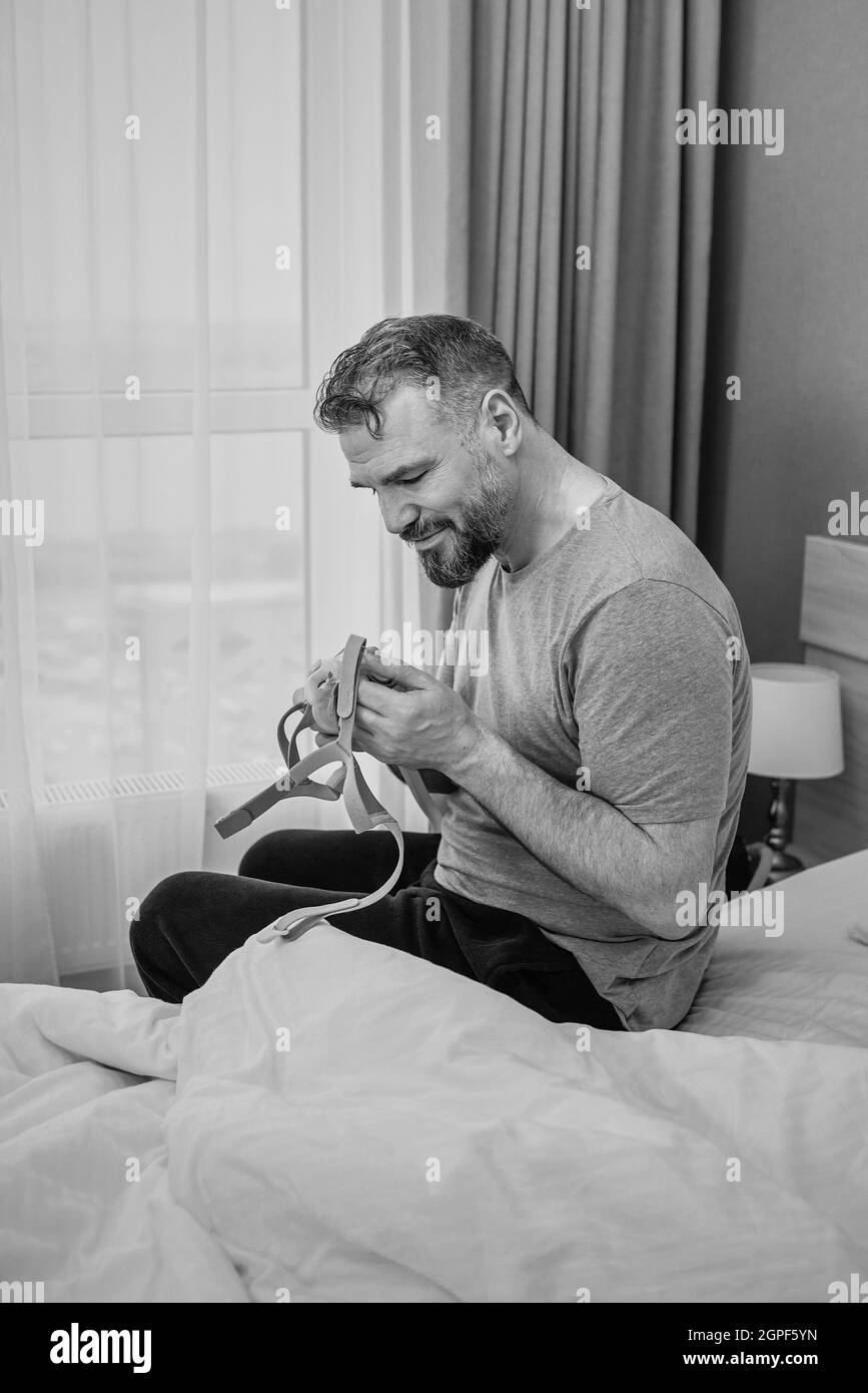 Reifer glücklicher Mann mit chronischen Atemproblemen erwägt die Verwendung CPAP-Maschine auf dem Bett im Schlafzimmer sitzen. Gesundheitswesen, CPAP, Schnarchen Konzept Stockfoto