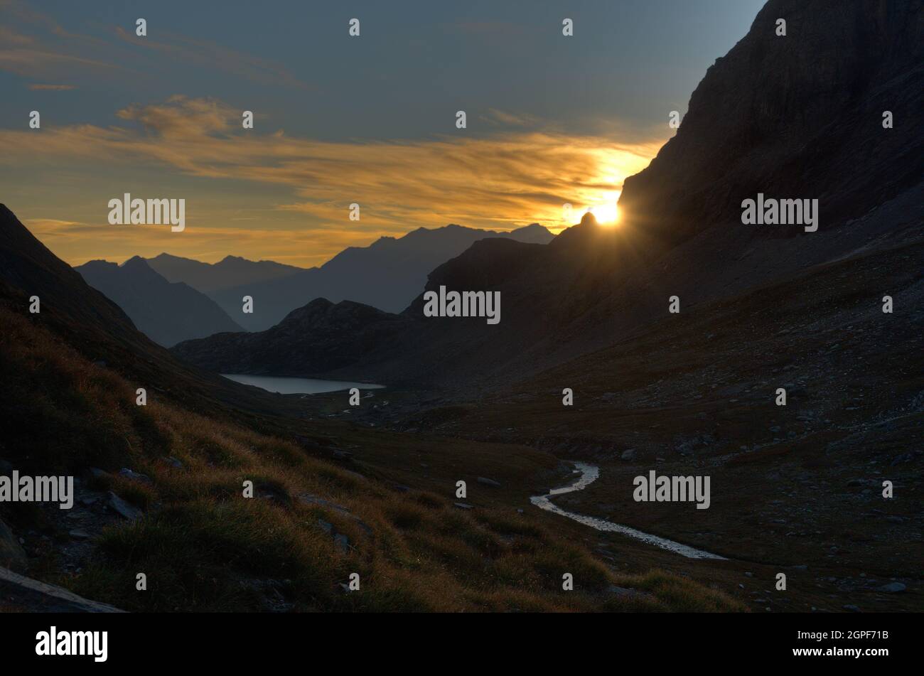 Am frühen Morgen in den Alpen: Silhouetten von Bergen in trüben Farben, ein silbriger See und ein Fluss im Vordergrund. Stockfoto