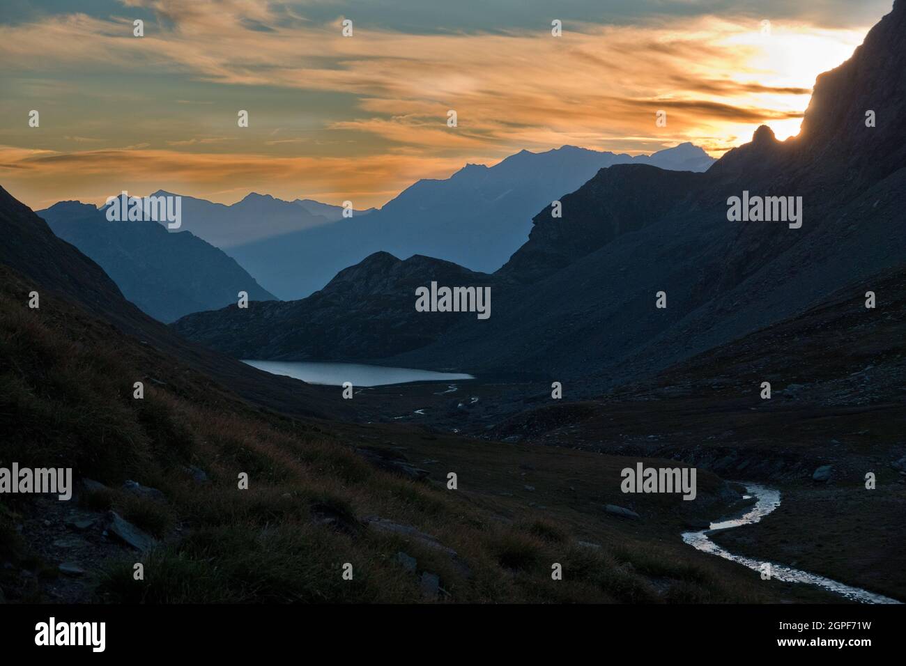 Am frühen Morgen in den Alpen: Silhouetten von Bergen in trüben Farben, ein silbriger See und ein Fluss im Vordergrund. Stockfoto