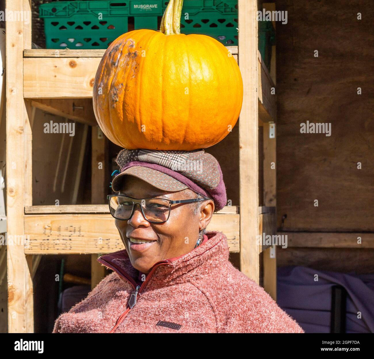 Ältere Frau, die Kürbisse verkauft, balanciert einen Kürbis auf ihrem Kopf an ihrem Marktstand aus der Nähe gegenüber der Kamera. Stockfoto