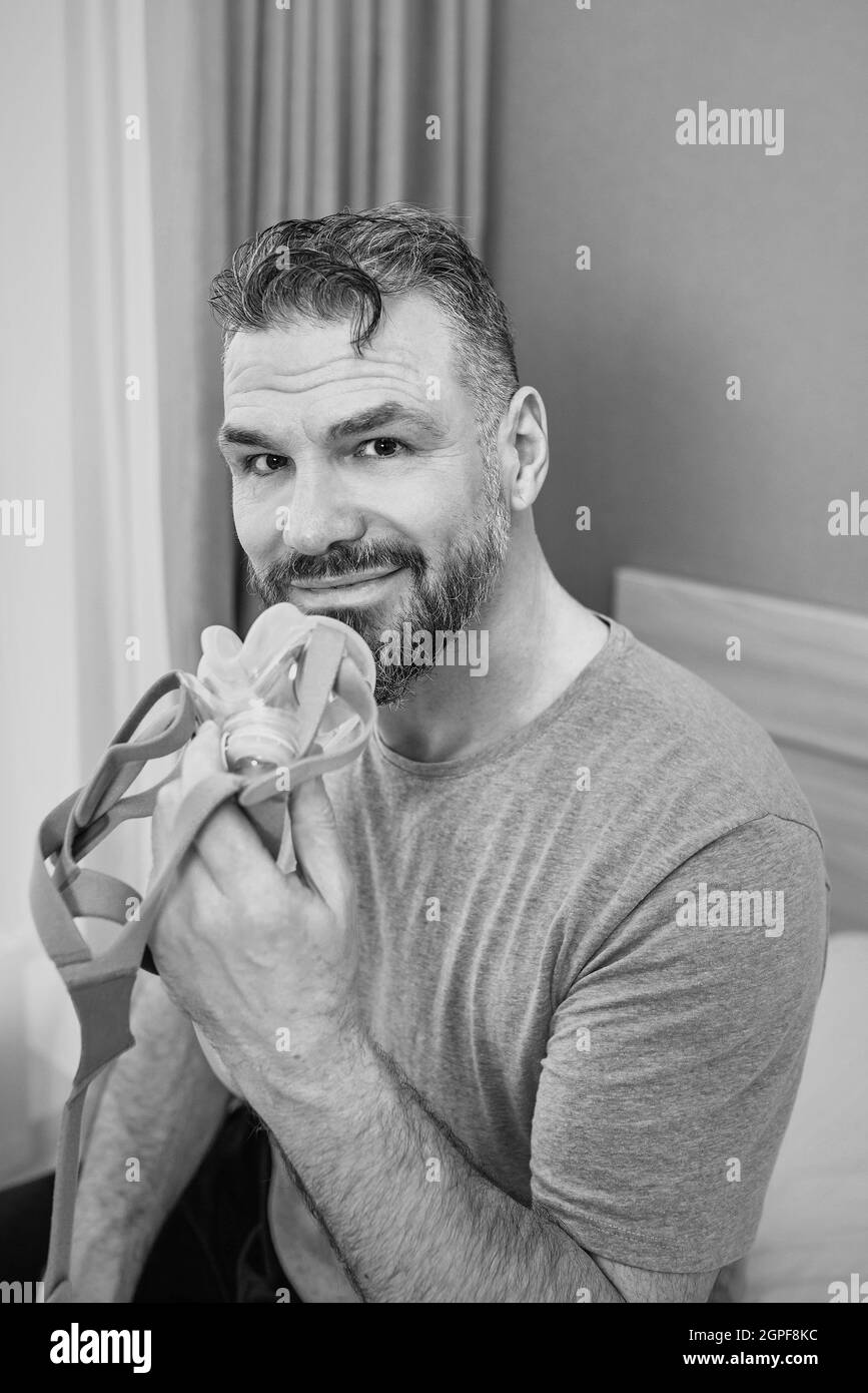 Reifer glücklicher Mann mit chronischen Atemproblemen erwägt die Verwendung CPAP-Maschine auf dem Bett im Schlafzimmer sitzen. Gesundheitswesen, CPAP, Schnarchen Konzept Stockfoto