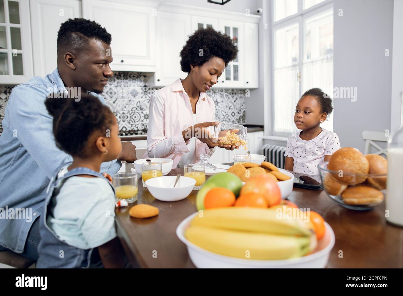 Hübsche afrikanische Schwestern, die mit jungen Eltern in einer modernen, hellen Küche sitzen. Positive Familie, die zum Frühstück Cerealien mit Milch isst. Morgens zu Hause. Stockfoto