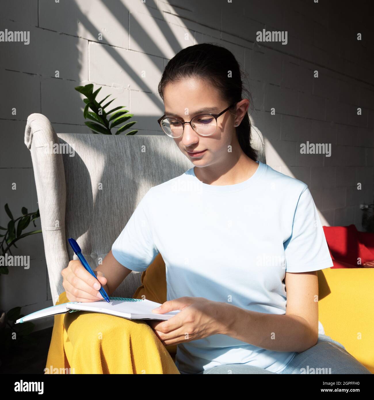 Eine hübsche Studentin schreibt mit einem Stift eine Aufgabe in einem Notizbuch, während sie in einem Café auf einem Sessel sitzt. Stockfoto