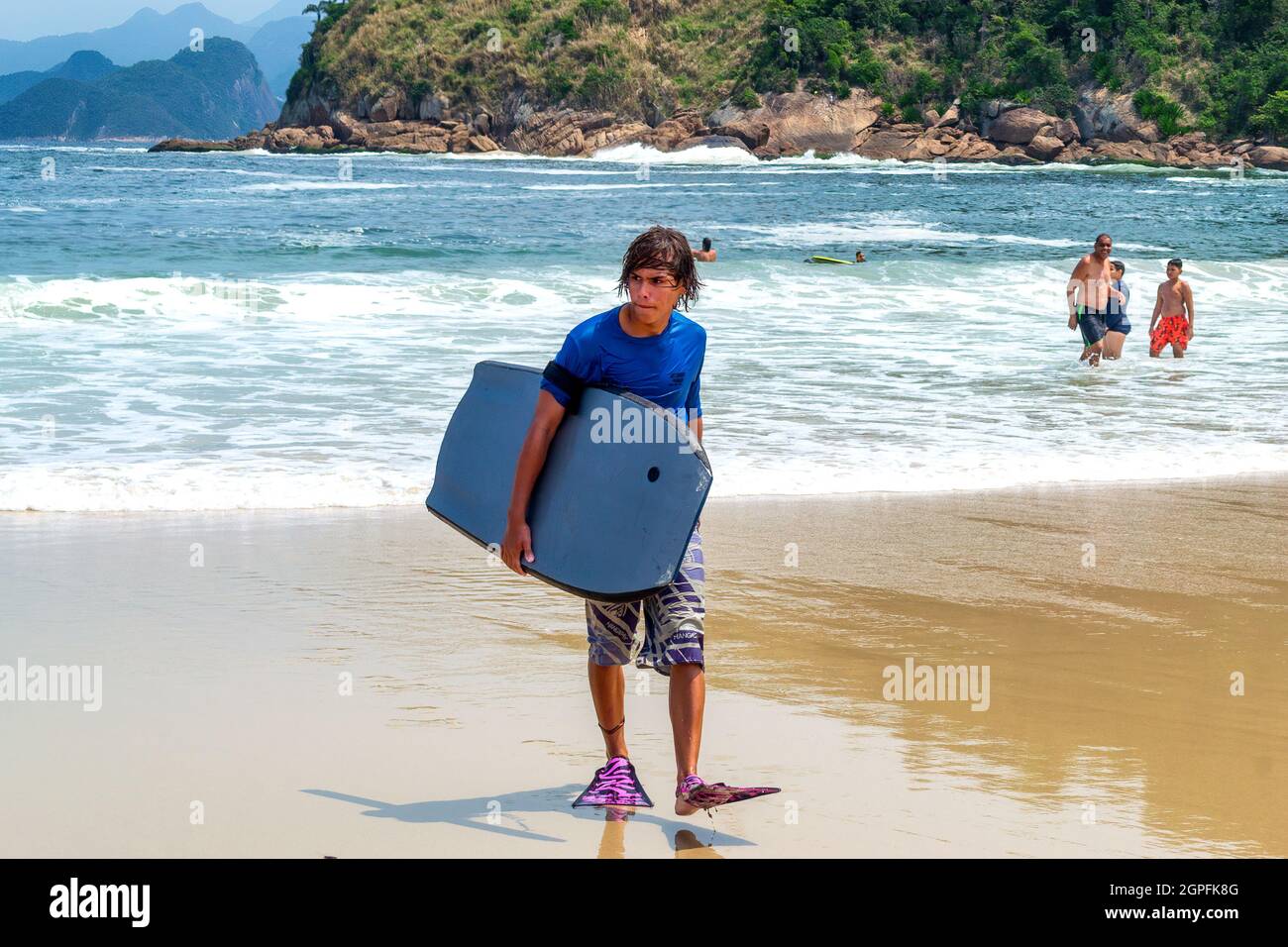 Brasilianischer junger Mann mit Surfbrett am Strand Piratininga in Niteroi, Rio de Janeiro, Brasilien. Der Wahrzeichen Strand ist ein berühmter Ort und ein Trave Stockfoto