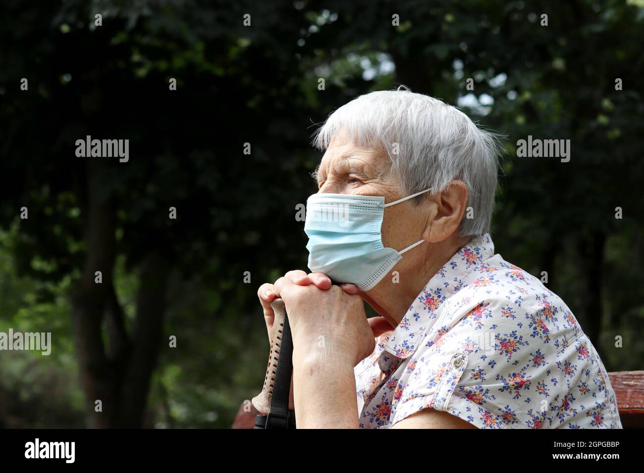Ältere Frau in Schutzmaske sitzt mit Gehstock auf einer Bank im Park. Sicherheit während einer Coronavirus-Pandemie, Leben im Ruhestand Stockfoto