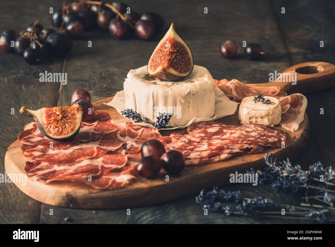 Verschiedene geräucherte Wurstspezialitäten und ein weicher Käse auf einem Holzbrett, verziert mit Feigen und Trauben Stockfoto
