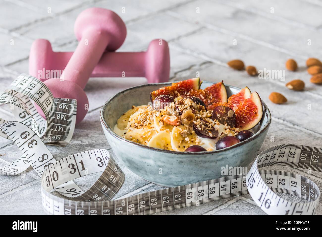 Schüssel mit Joghurt, Obst und Nüssen für eine gesunde Ernährung Frühstück oder Snack. Rosa Kurzhanteln und ein Maßband auf grauem Hintergrund, Gewichtsverlust Konzept Stockfoto