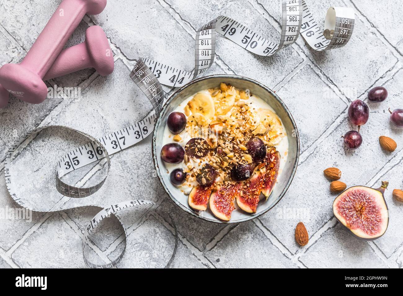 Schüssel mit Joghurt, Obst und Nüssen für eine gesunde Ernährung Frühstück oder Snack. Rosa Kurzhanteln und ein Maßband auf grauem Hintergrund, Gewichtsverlust Konzept. Oben Stockfoto