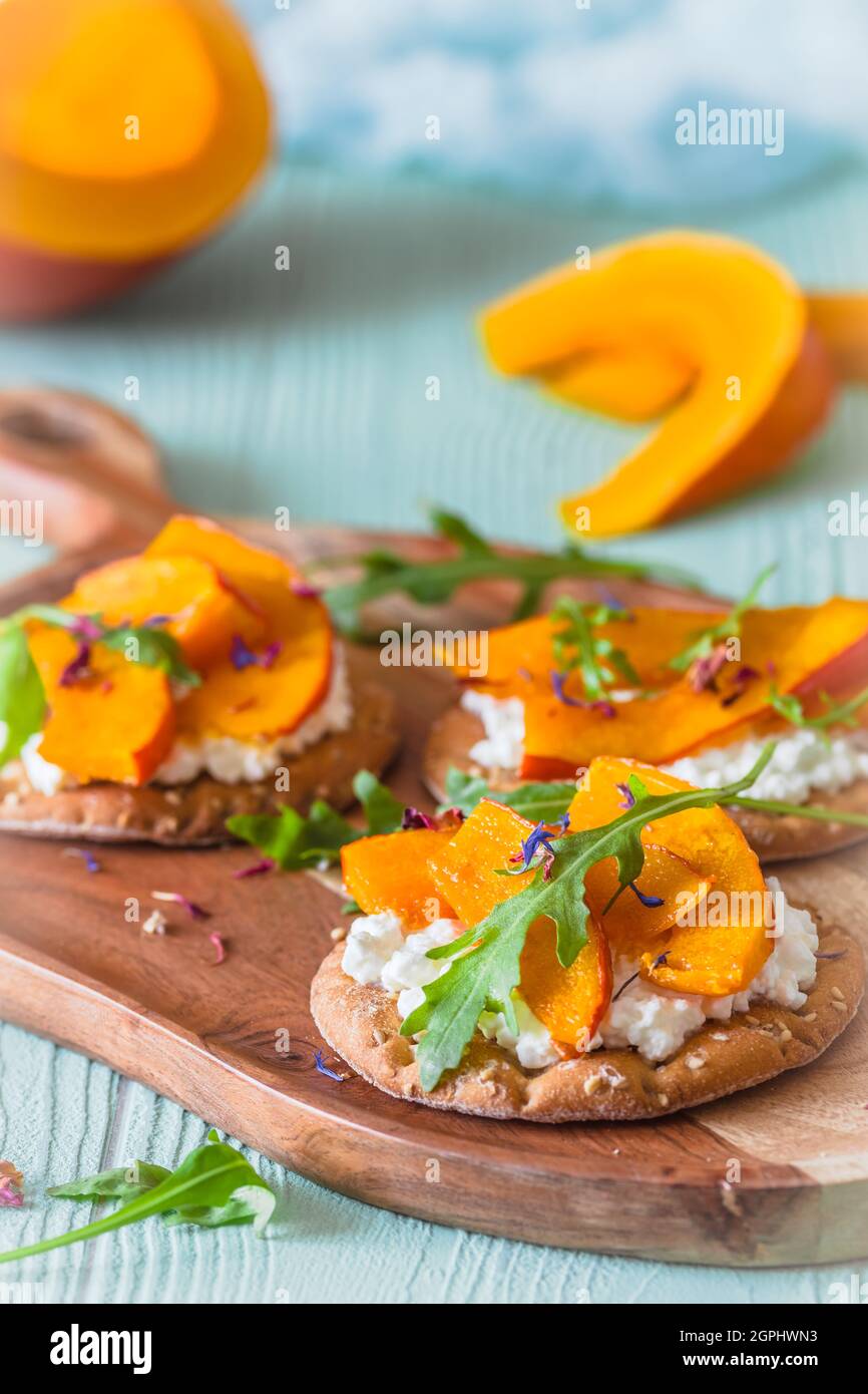 Runde Knäckebrot-Scheiben mit geröstetem Kürbis, körnigem Frischkäse und Rucola auf einem Holzbrett auf hellgrünem Hintergrund, vertikal Stockfoto