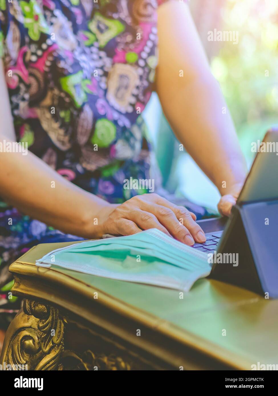 Hände einer reifen Frau, die auf der Laptop-Tastatur tippt, um von zu Hause aus zu arbeiten und ihre OP-Maske auszuziehen, legen sie auf den Tisch, damit sie leichter atmen kann. Konzepte von C Stockfoto