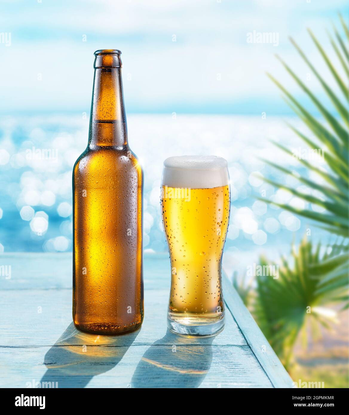 Geöffnete Flasche Bier und Bierglas mit Kondenswasser auf dem Holztisch. Verschwommenes glitzerndes Meer im Hintergrund. Sommerliche Stimmung. Stockfoto