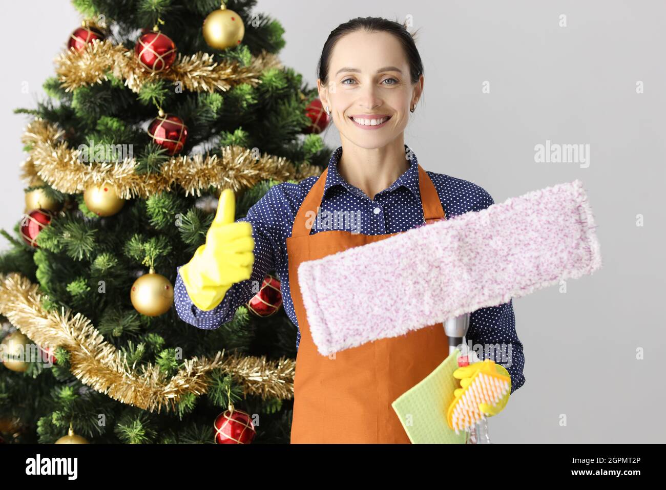 Porträt einer jungen lächelnden Frau, die die Daumen mit Mopp und Eimer auf dem Hintergrund des Weihnachtsbaums hochhält Stockfoto