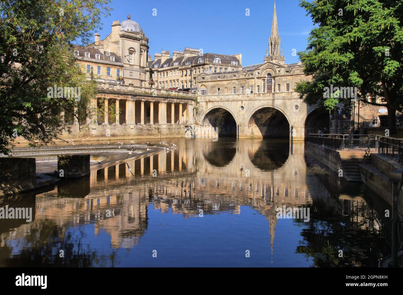 Kolonnaden, Kirche, Wehr und Pulteney Bridge über den Fluss Avon mit Reflexionen kurz nach Sonnenaufgang in Bath, Somerset, England Stockfoto