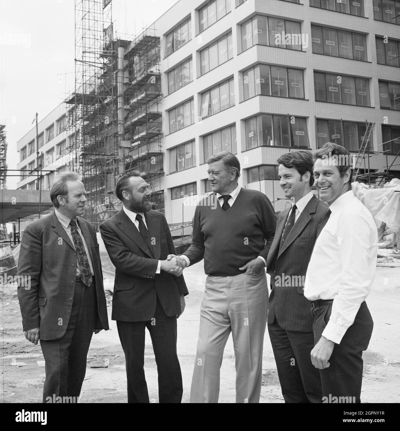 Der Schauspieler John Wayne trifft Mitarbeiter, die auf der Baustelle des St. Thomas' Hospital arbeiten. Dieses Foto wurde während der Dreharbeiten von Brannigan unter der Regie von Richard Attenborough aufgenommen. Die Aufnahmen wurden im St. Thomas' Hospital und John Wayne zusammen mit seiner Co-Star Judy Geeson gedreht, und der Regisseur machte sich mit dem Ort vertraut. Das Foto wurde auch im September 1974 im monatlichen Newsletter „Team Spirit“ von Laing veröffentlicht. Es zeigt verschiedene Mitarbeiter, die am Laing-Bauvertrag im Krankenhaus gearbeitet haben, und John Wayne wird gesehen, wie er dem Projektleiter D Upton die Hand schüttelte. Stockfoto