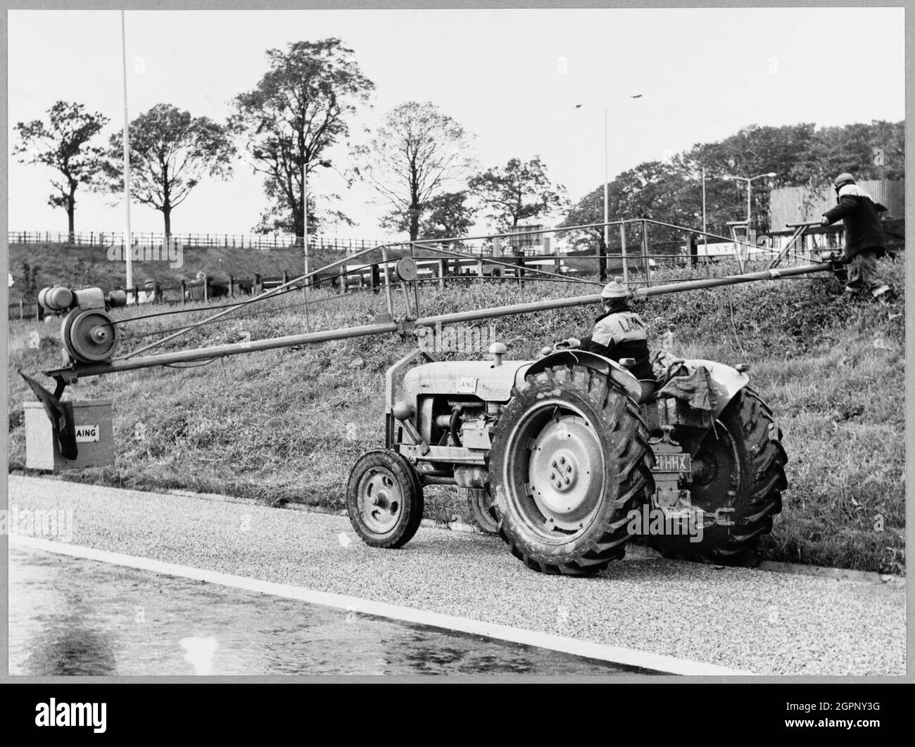 Zwei Laing-Arbeiter schneiden Gras auf einem Abschnitt der Autobahn Birmingham nach Preston (M6) zwischen Junction 13 und Junction 16, wobei ein Traktor mit Anbaugerät für die Auslegermaschine verwendet wird. Dieses Bild wurde als Teil des Breaking New Ground Project in Zusammenarbeit mit dem John Laing Charitable Trust in den Jahren 2019-20 katalogisiert. Stockfoto