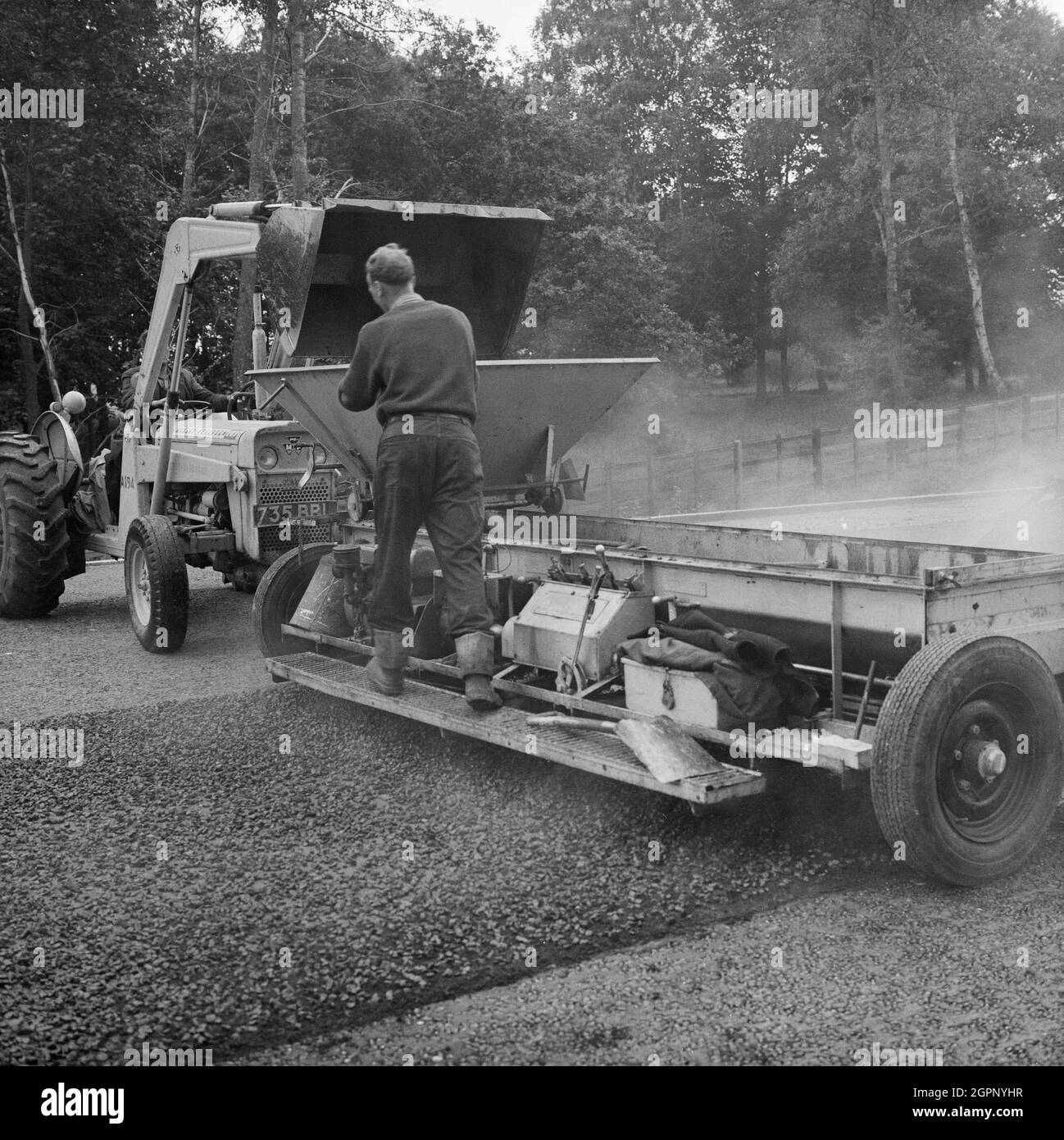 Nahaufnahme eines Splitterstreuers, der während des Baus der Birmingham to Preston Motorway (M6) in Betrieb war, mit einem Mann, der hinten stand und beobachtete, wie ein Traktor das Aggregat in den Trichter auslaste. Dieses Bild wurde als Teil des Breaking New Ground Project in Zusammenarbeit mit dem John Laing Charitable Trust in den Jahren 2019-20 katalogisiert. Stockfoto