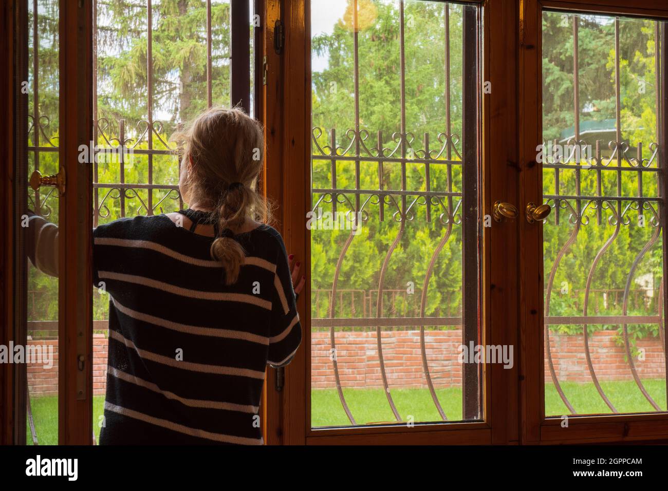 Die junge Frau beobachtet den Garten ihres Hauses durch die verbarsten Holzfenster. Stockfoto
