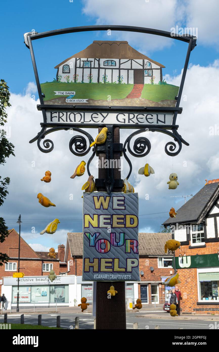 Frimley Green Village Schild mit gestrickten oder genähten Kanarienvögel, die von einer sanften Klimaaktivismus-Bewegung namens canary Crafttivists, Surrey, Großbritannien, daran hängen Stockfoto