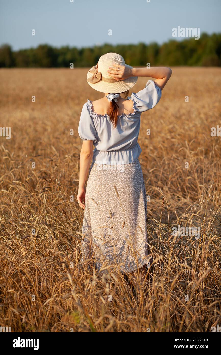 Junge schöne Mädchen mit langen lockigen Haaren Posen in einem Weizenfeld im Sommer bei Sonnenuntergang. Ein Mädchen hält einen Hut in der Hand vor dem Hintergrund eines W Stockfoto