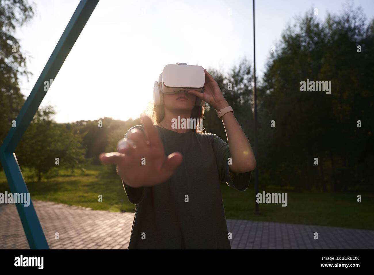 Nahaufnahme eines Mädchens, das mit den Händen eine Virtual-Reality-Brille auf dem Kopf hält und auf einem Hintergrund von grünen Bäumen steht. Er reicht eine Hand an die Kamera. Hochwertige Fotos Stockfoto