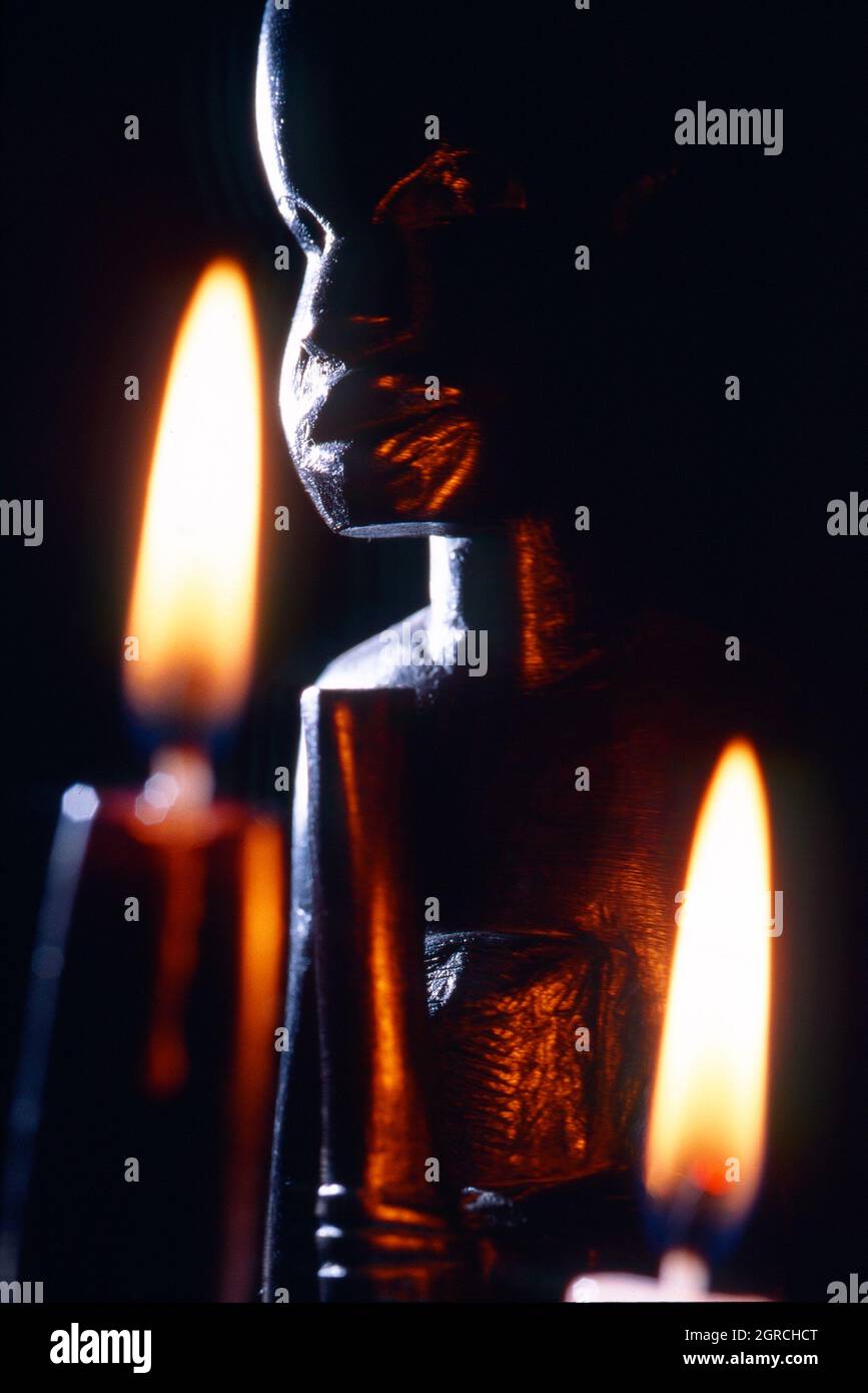 Dunkle Nahaufnahme einer afrikanischen Statue mit zwei brennenden Kerzen, einer schwarz und der andere weiß Stockfoto