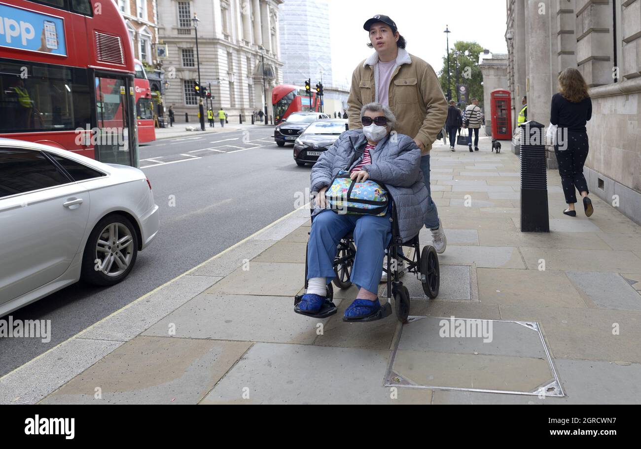 London, England, Großbritannien. Mann, der eine Frau mit Gesichtsbefrag in einen Rollstuhl schiebt, 2021 Stockfoto
